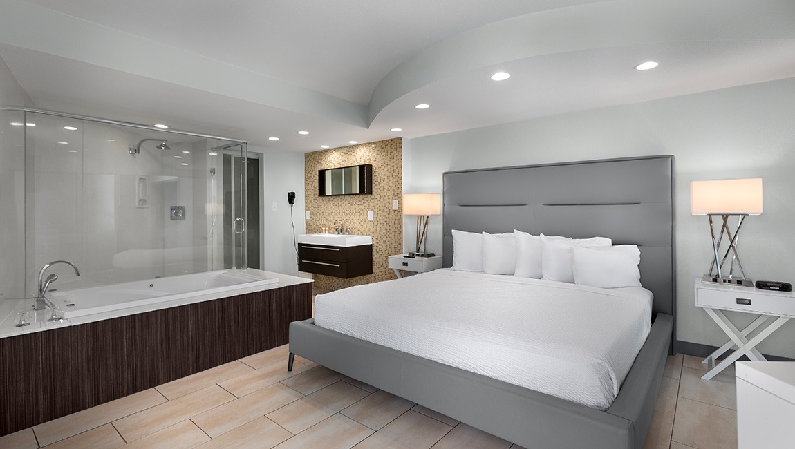 A large hotel room with a gray bed, a jacuzzi tub, and a glass-enclosed shower.