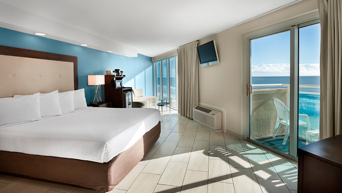 A hotel room with a large bed, a sofa, a TV, and a balcony with an ocean view.