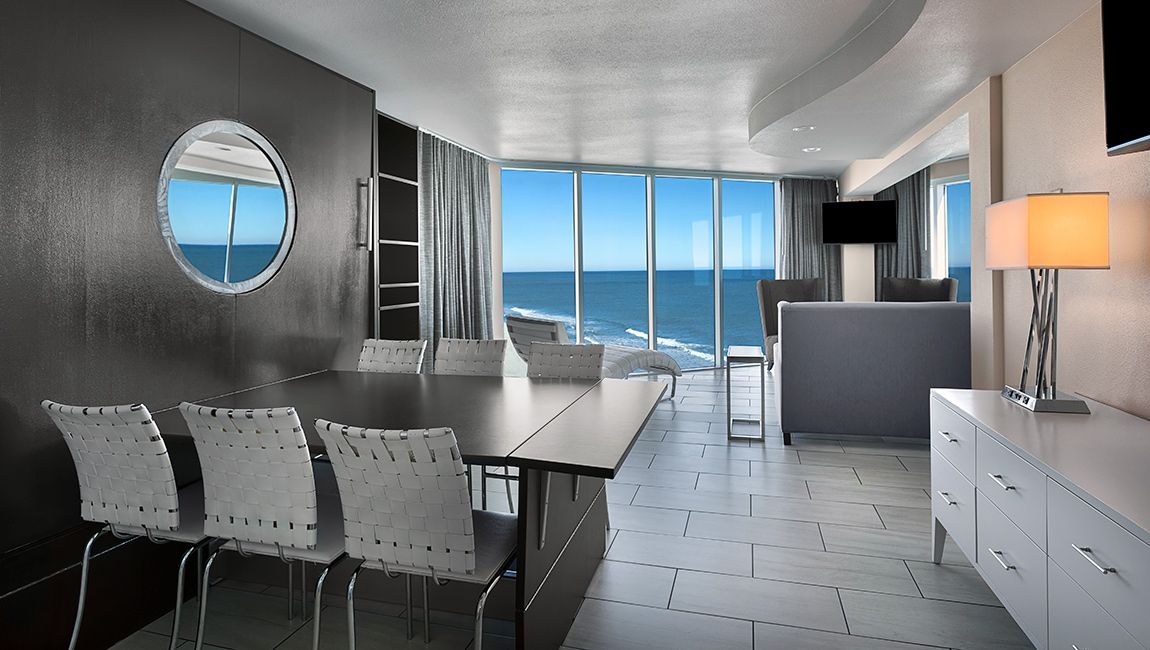 A wide view of a modern room with a dining table, sofa, and large windows showing the ocean.