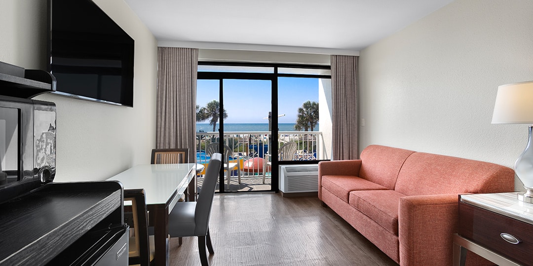 A wide view of a hotel suite with a sofa, dining table, and balcony with an ocean view.
