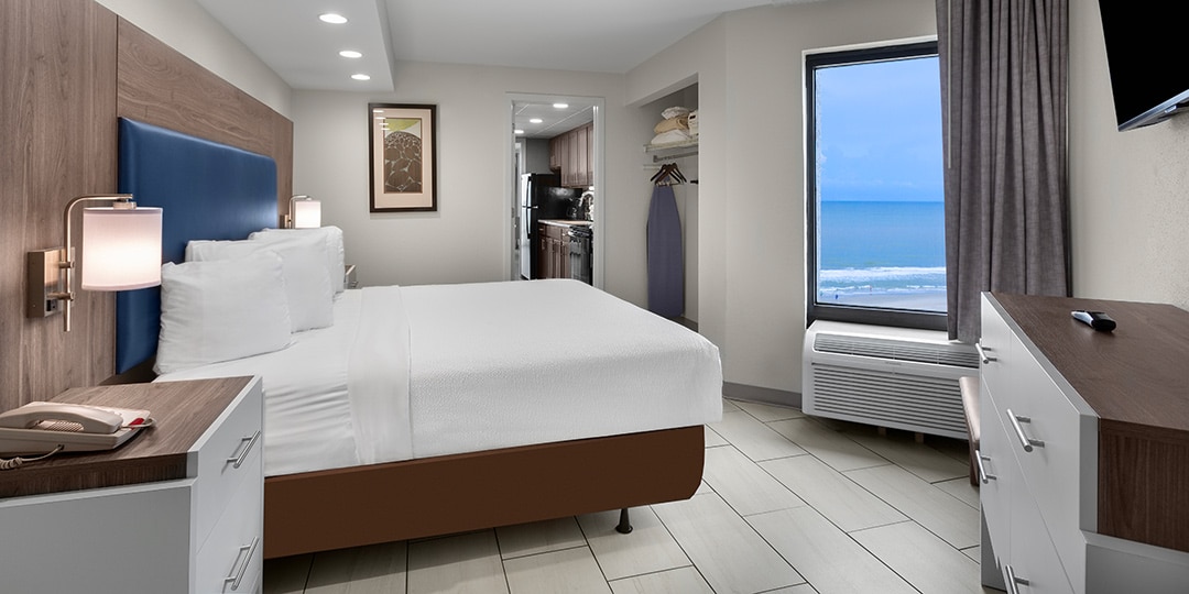A wide view of a hotel room with a king-size bed, ocean view, kitchenette, closet, and flat-screen TV.