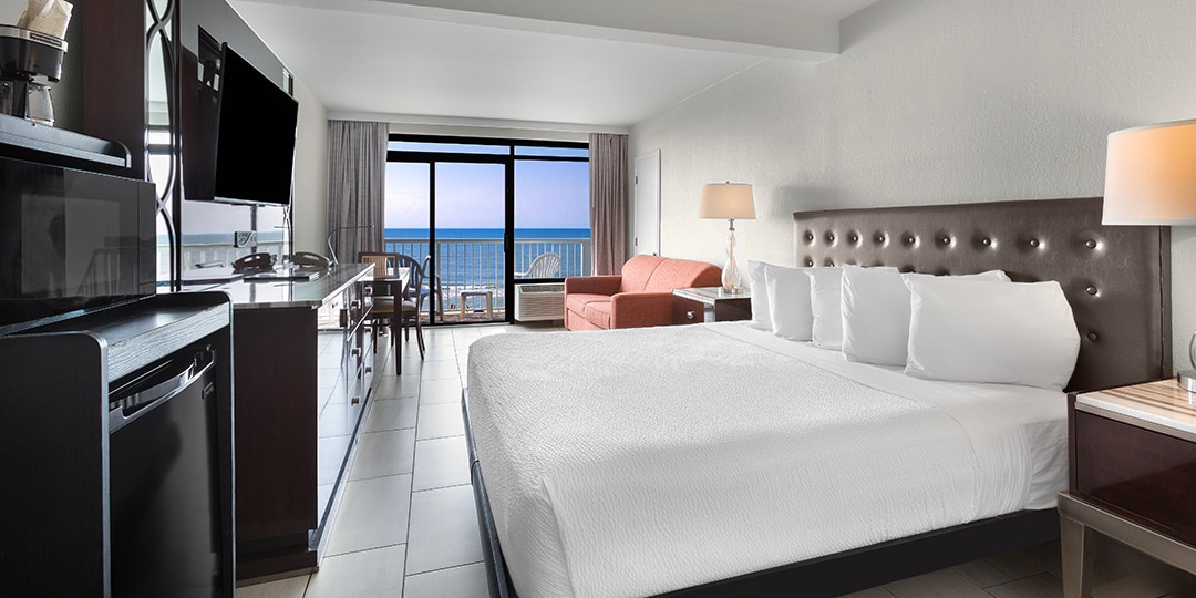 A wide view of a hotel room with a large bed, kitchenette, and balcony with ocean view.