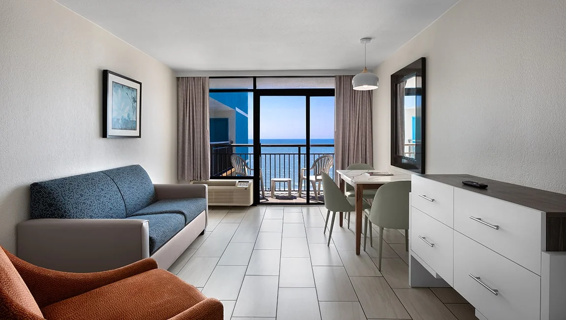 A wide view of a hotel room with a sofa, dining table, and balcony with an ocean view.