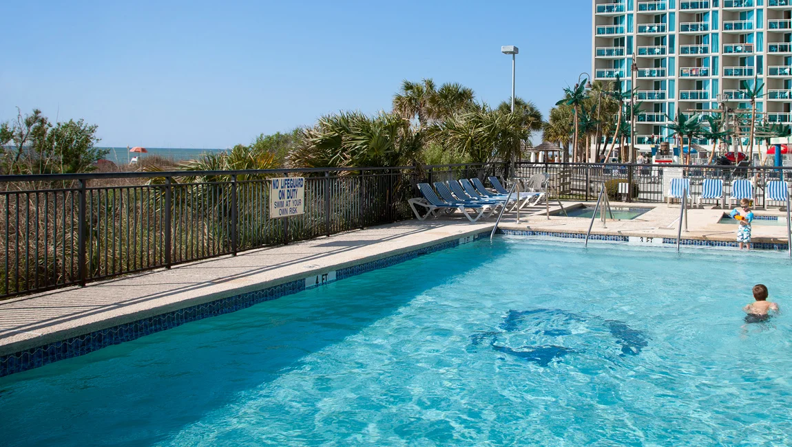 A fenced outdoor pool with blue water and lounge chairs, surrounded by palm trees and a high-rise building in the background.
