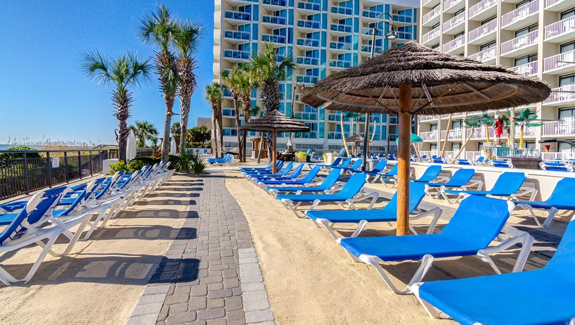 A resort pool area with blue lounge chairs, thatched umbrellas, and palm trees, in front of a high-rise building.